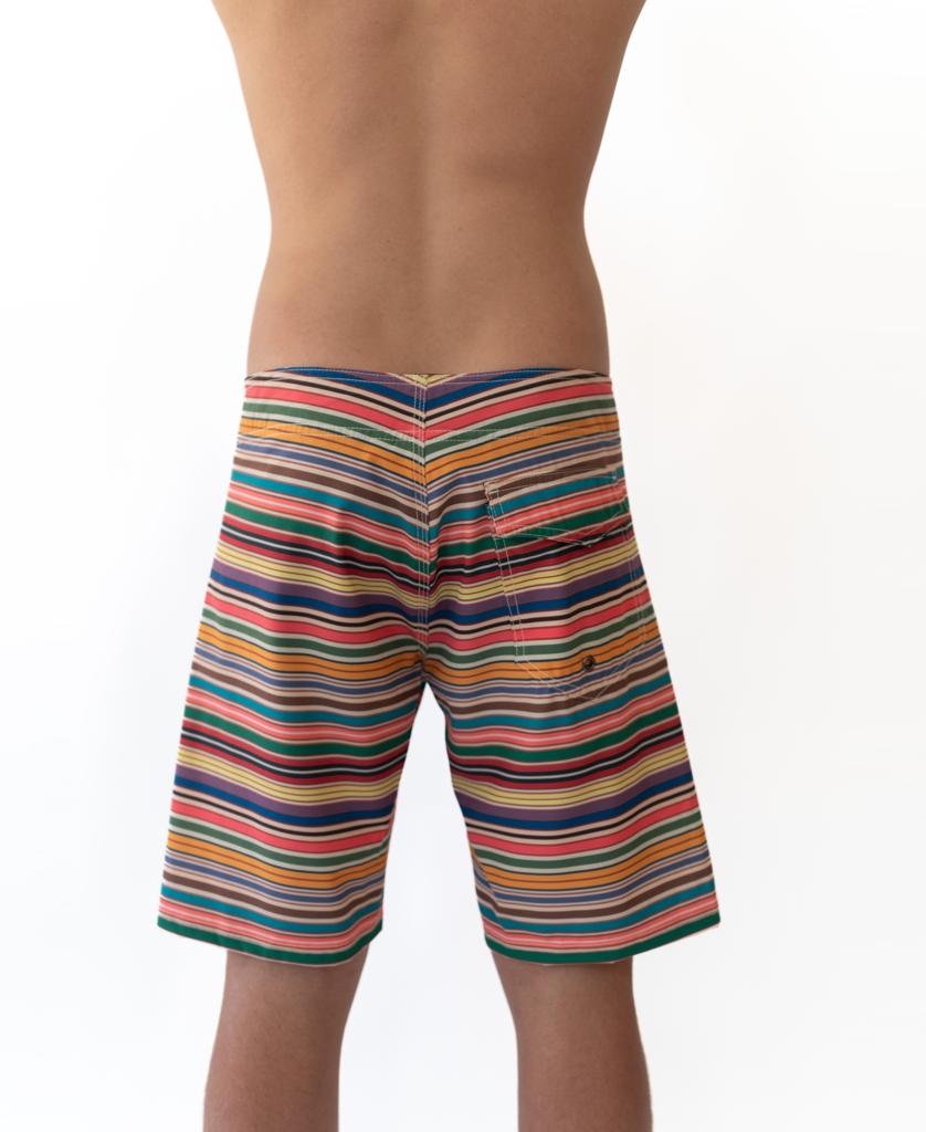 A person stands facing away, wearing Golden Breed’s Daze Boardie | Stripe—multicolored striped boardshorts with a back pocket, made from recycled polyester—set against a plain white background.