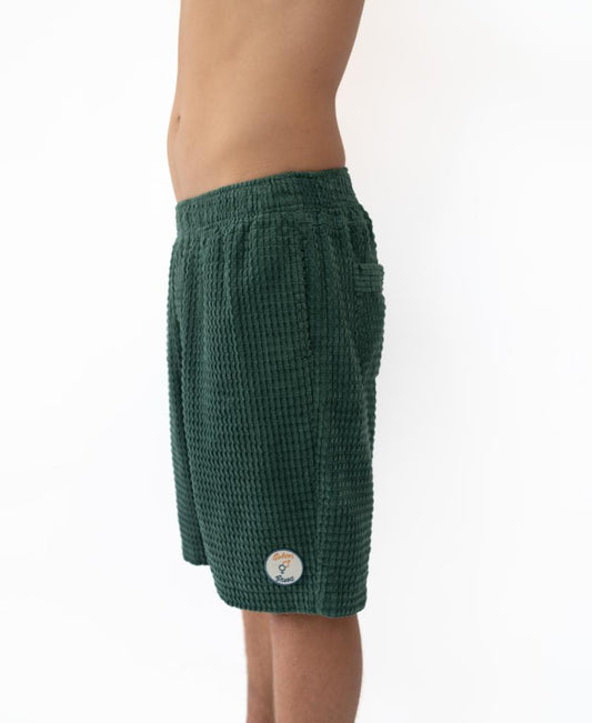 A person stands in profile against a plain white background, wearing Golden Breed’s El Solito Walkshort in Ivy Green, made from chunky cotton cord and featuring a small circular patch above the left hem.