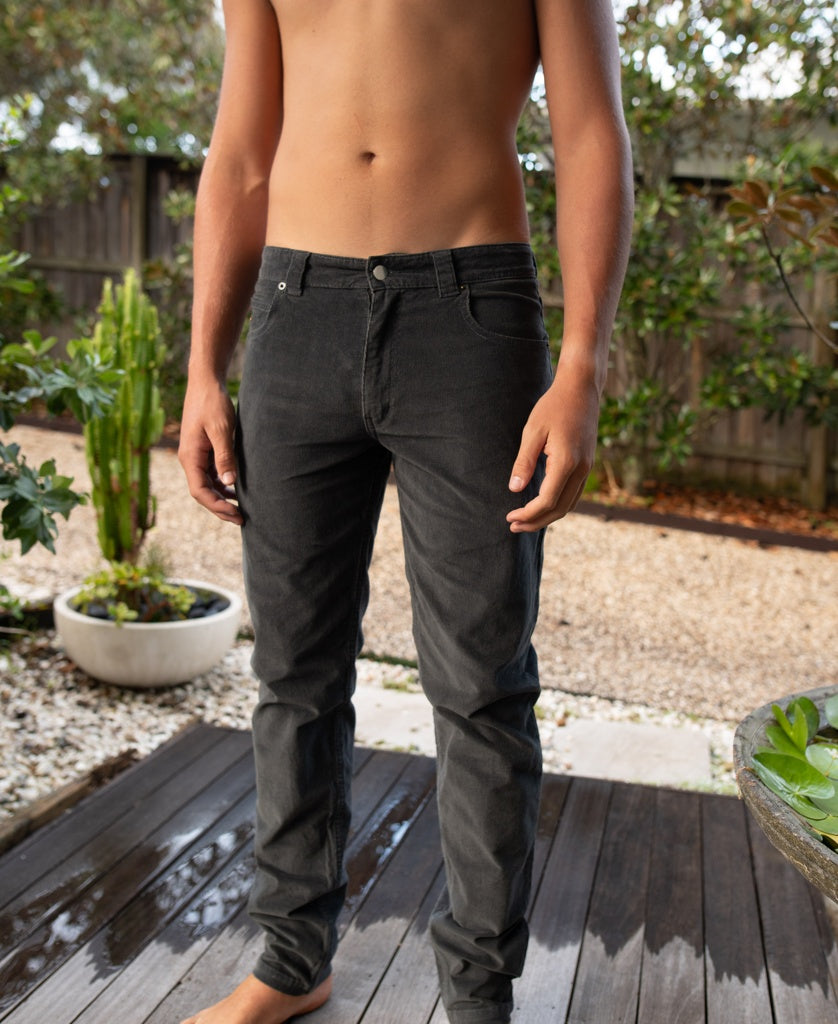 A shirtless person stands outdoors on a wooden deck, wearing Golden Breed Felix Stretch Cord Pant in wash black, with plants and a gravel area visible in the background.