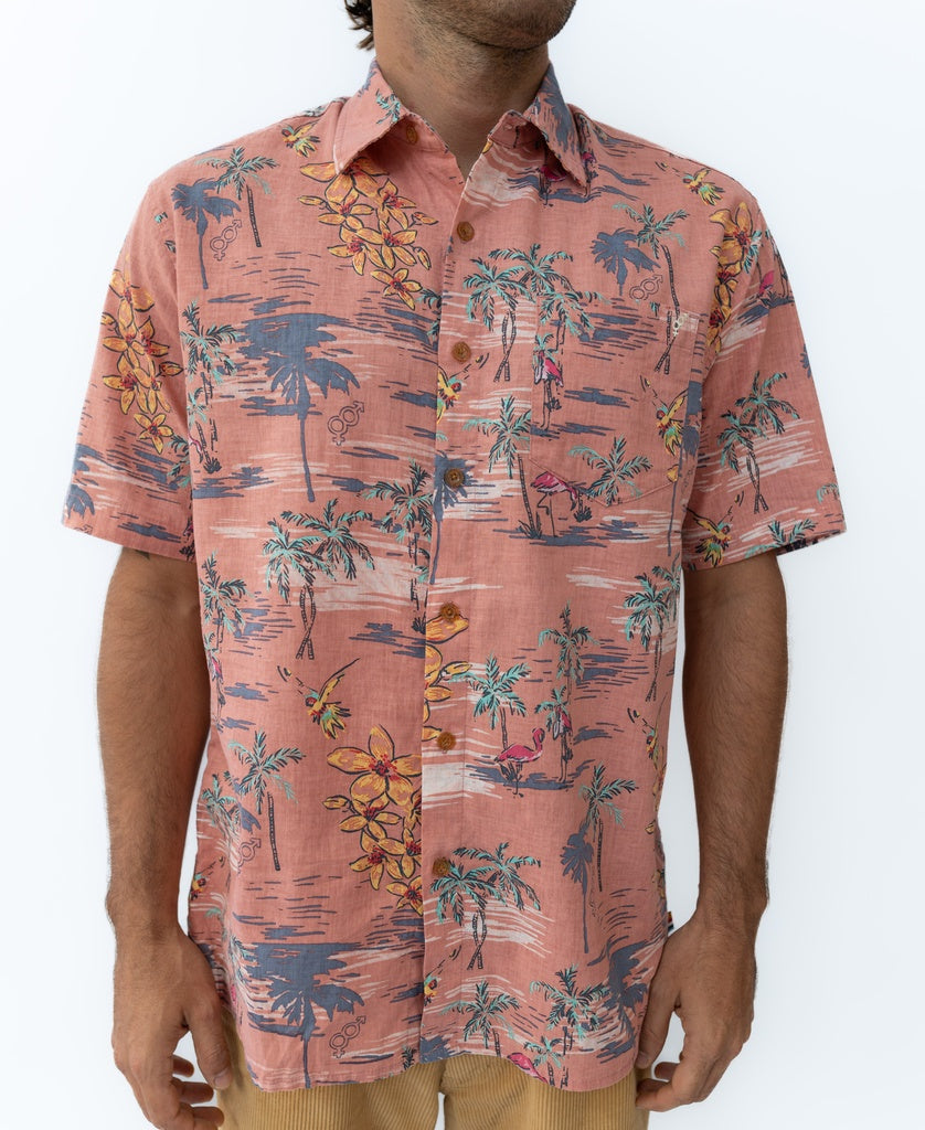 A man wears the Golden Breed Flamingos Shirt | Rose, a short-sleeve button-up made from a breathable linen blend with palm trees, flowers, and people, standing against a plain white background.
