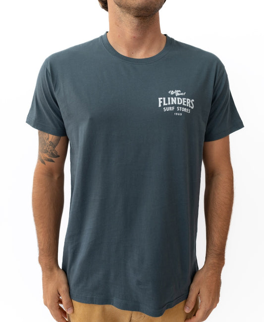 A man wears the Golden Breed Flinders Easy Rider Tee in Denim, made from 100% Cotton Jersey with FLINDERS SURF STORES 1968 on the left chest, standing against a white background.