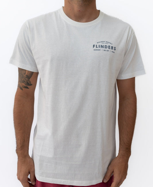 A person wears the Golden Breed Flinders Old School Tee in off white, a 100% cotton shirt featuring a small FLINDERS logo on the upper left chest, paired with red shorts. Only their upper body is visible.