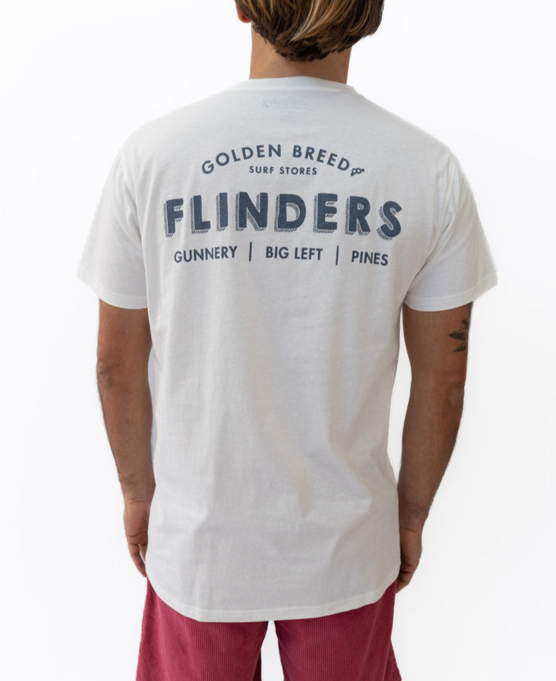 A person wearing a Golden Breed Flinders Old School Tee in Off White with the Flinders Gunnery Big Left Pines print on the back, paired with red shorts.