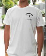 A person wearing the Golden Breed Flinders Shanty Tee in Off White, featuring a small left chest graphic, stands outdoors near a surf store.