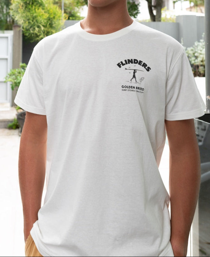 A person wearing the Golden Breed Flinders Shanty Tee in Off White, featuring a small left chest graphic, stands outdoors near a surf store.