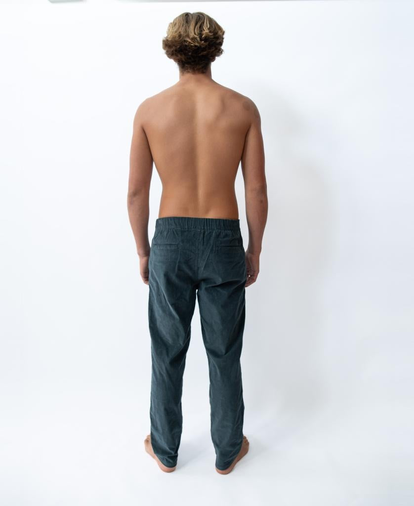 A shirtless person with short, light brown hair stands barefoot facing away from the camera, wearing Golden Breeds Fred Stretch Cord Pant in blue against a plain white background.