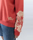 A person wears the Golden Breed Hibiscus Sweat in Wash Red/Beige, made from 100% cotton fleece with bold text and a large floral sleeve graphic, paired with blue denim shorts.
