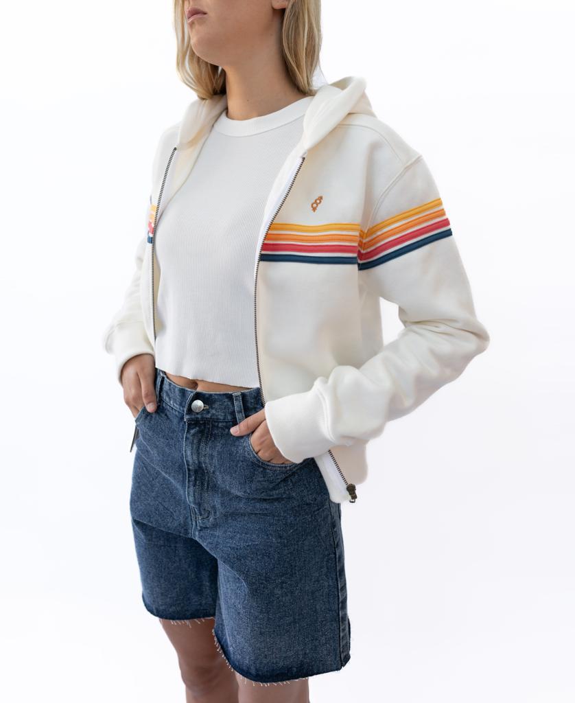 A person stands with hands in pockets against a white background, wearing Golden Breeds Izzy Short in Dark Blue, paired with a white hoodie featuring orange and blue stripes over a white cropped top.