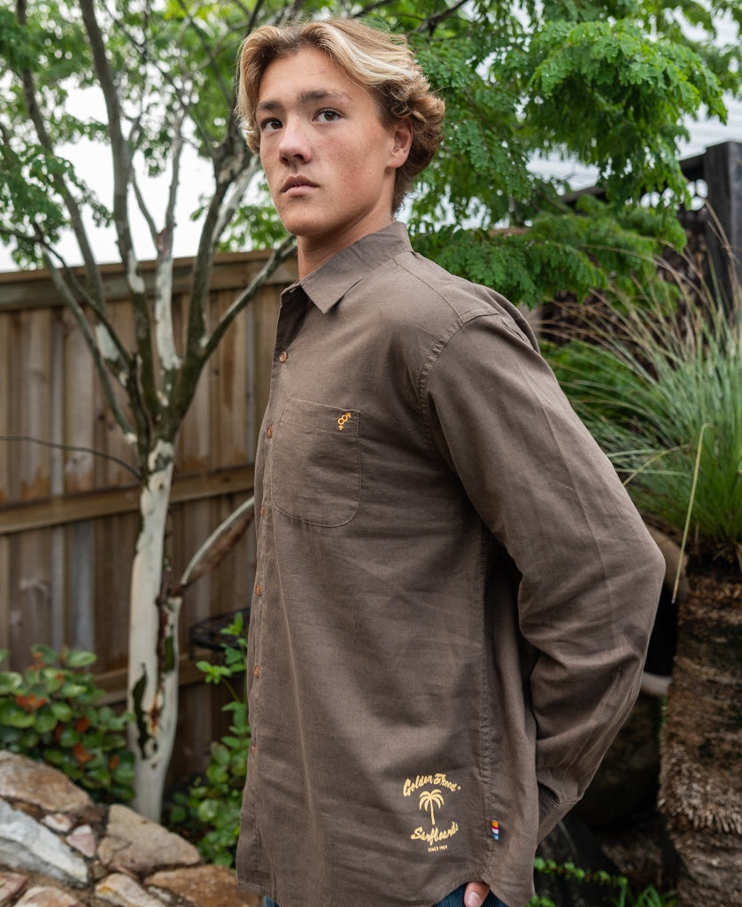 A young man with blonde hair wears the Golden Breed Linen LS Shirt in Wash Brown, standing outside by a wooden fence amid lush garden greenery.