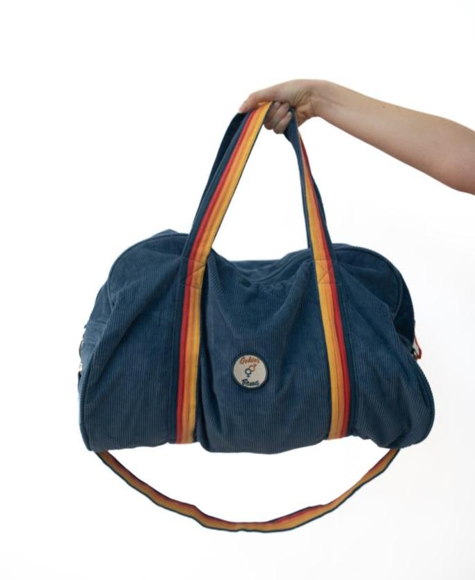 A hand holds the Golden Breed Long Island Cord Roll Bag in Dusty Blue, featuring rainbow-striped handles and a round front patch—an ideal weekend getaway bag—set against a white background.