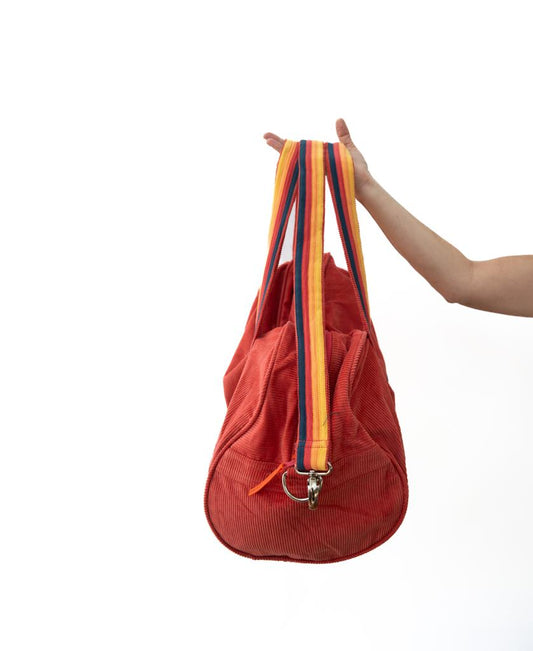 A person holds up a Golden Breed Long Island Cord Roll Bag in Wash Red, featuring multicolored striped cotton cord handles, against a plain white background.