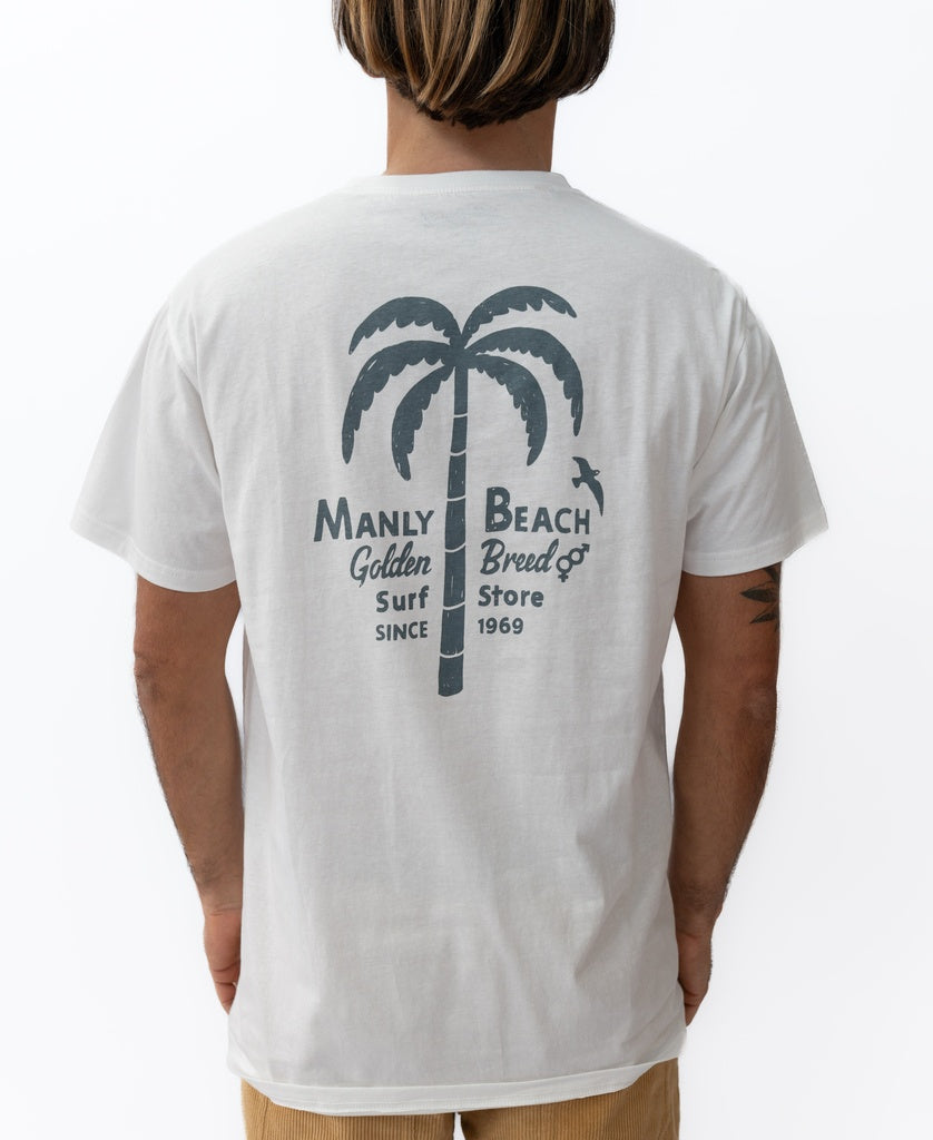 A person wearing the Manly Palm Loco Tee by Golden Breed, an off-white 100% cotton T-shirt featuring a palm tree graphic and “Manly Beach Golden Breed Surf Store Since 1969” on the back. Designed in Australia.