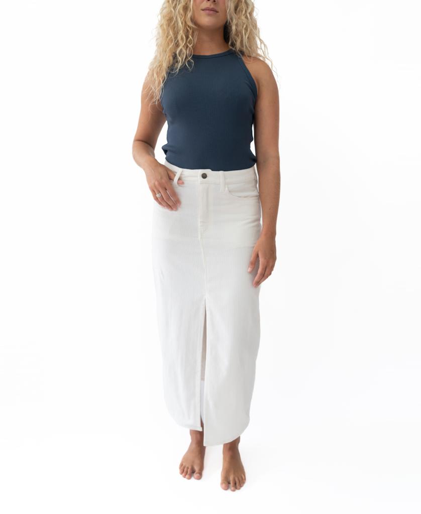A person with long curly hair wears a navy sleeveless top and the Nellie Maxi in Off White by Golden Breed, featuring a high waist and front slit, while standing barefoot against a plain white background.