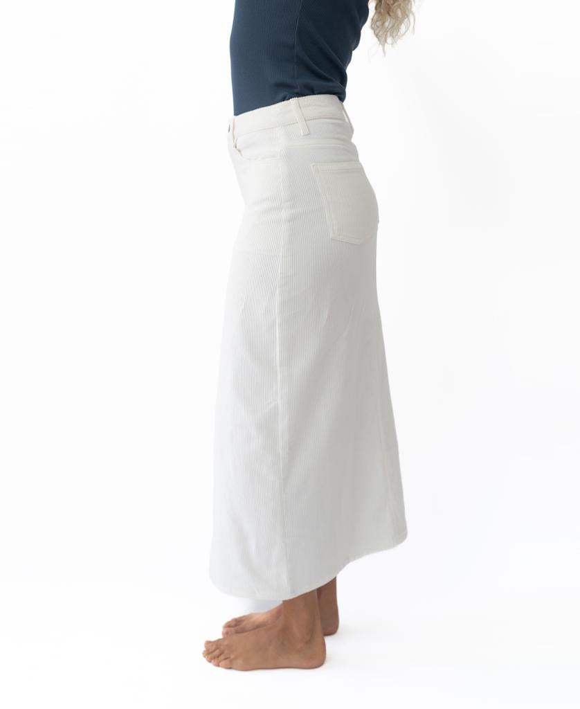 A person in a dark top and the Golden Breed Nellie Maxi | Off White skirt stands barefoot on a white background, shown from the side. This elegant skirt is handmade in Indonesia from premium cord fabric.