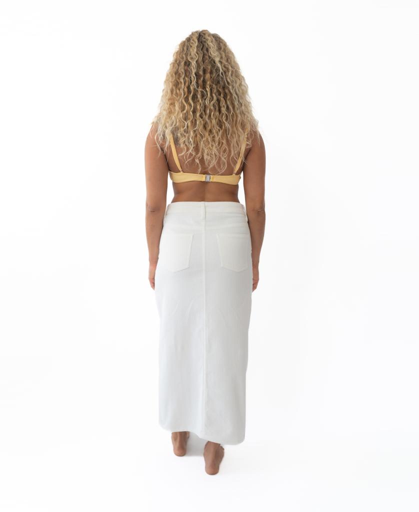 A person with long curly hair, wearing a yellow top and the Golden Breed Nellie Maxi in Off White, stands barefoot facing away against a white background.