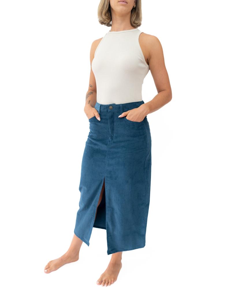 A woman stands barefoot with hands in pockets, wearing a sleeveless white top and the Golden Breed Nellie Maxi Skirt in Petrol—a blue cord fabric skirt with a front slit.