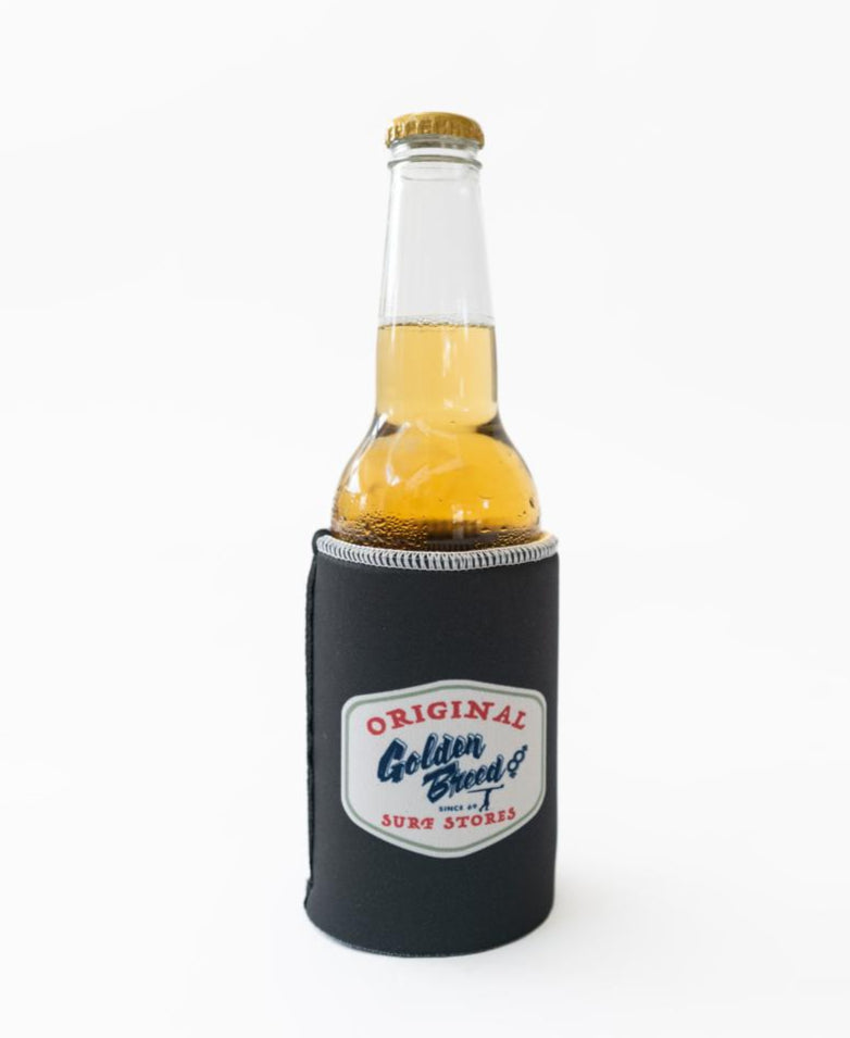 A Golden Breed Ray Stubby Holder in wash black holds a clear glass bottle of light beer; the stubby holder features Original Golden Breed Surf Stores branding on a white background.