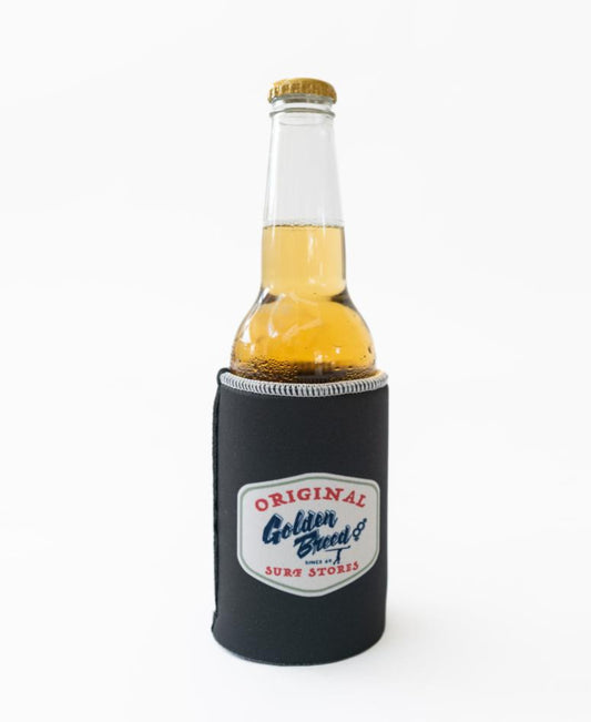 A Golden Breed Ray Stubby Holder in wash black holds a clear glass bottle of light beer; the stubby holder features Original Golden Breed Surf Stores branding on a white background.
