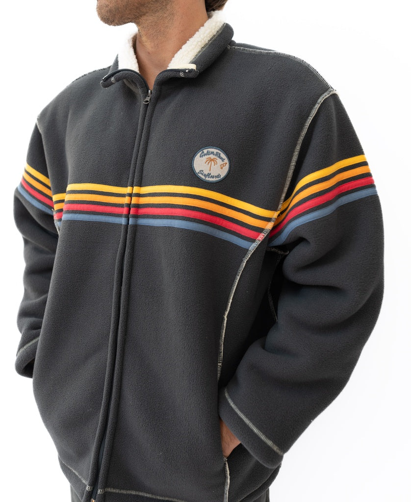 A person wearing the Golden Breed Reef Bonded Sherpa Jacket in Wash Black, featuring red, yellow, and blue stripes and a round chest patch, made from cozy polyester sherpa.
