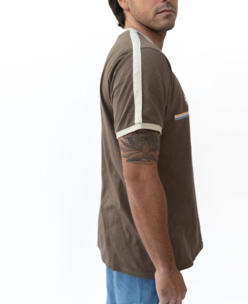 A man wearing the Golden Breed Retro Ringer Tee in Wash Brown with white sleeve stripes stands sideways, displaying a tattoo on his upper arm against a white backdrop, reflecting classic 70’s style.