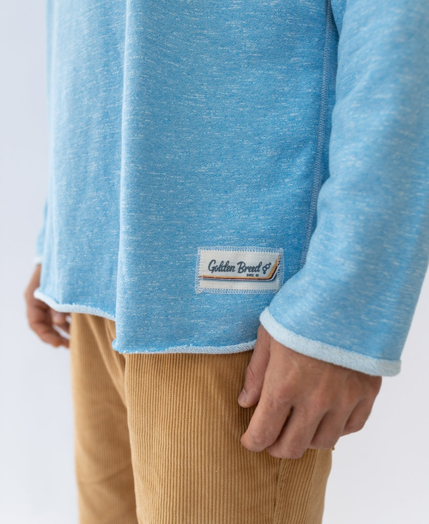 A person wears the Golden Breed Retro Rag Top in Sky Marle—a light blue long-sleeve with an embroidered GB logo and label—paired with tan corduroy pants in a slouchy, retro-inspired style, shown shoulders down.