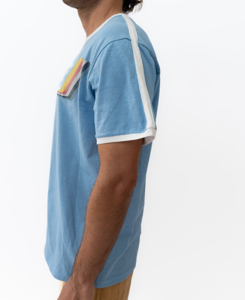 A man wears the Golden Breed Retro Ringer Tee | Azure Blue, featuring white trim and a rainbow-striped chest patch, standing against a plain white background for classic 70s style.