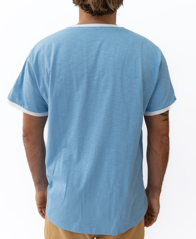 A person stands with their back to the camera, wearing Golden Breed’s Retro Ringer Tee in Azure Blue with white trim and light brown pants, channeling effortless 70s vibes.