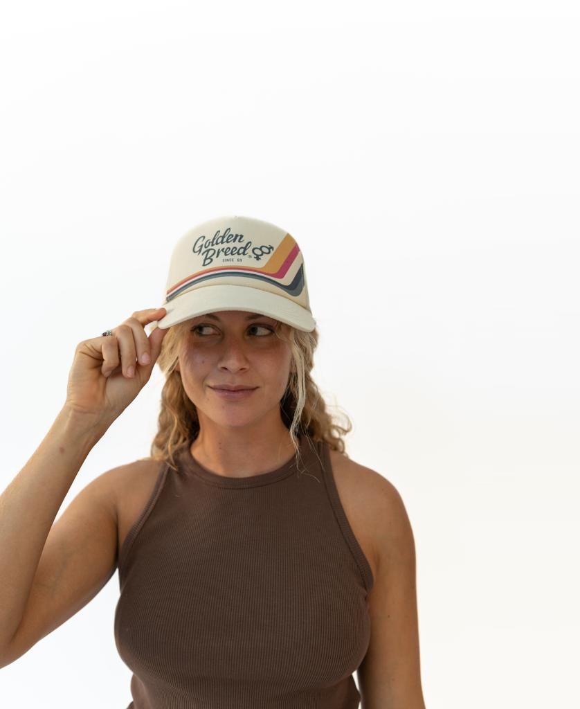 A woman wears a brown sleeveless top and an off white Retro Trucker Cap by Golden Breed, featuring an adjustable strap and rainbow stripe, standing against a plain white background.