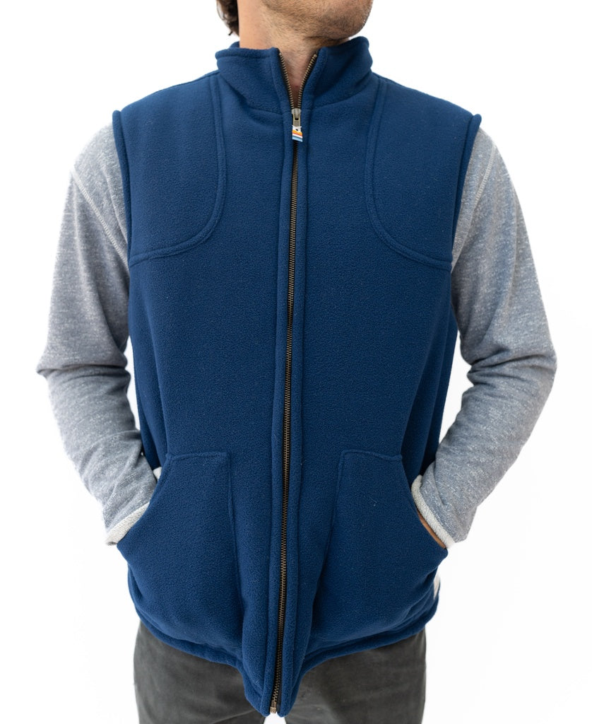A person wears a sleeveless Golden Breed Thredbo Vest | Midnight, made from polyester polar fleece, over a gray long-sleeve shirt with hands in vest pockets, standing against a white background.