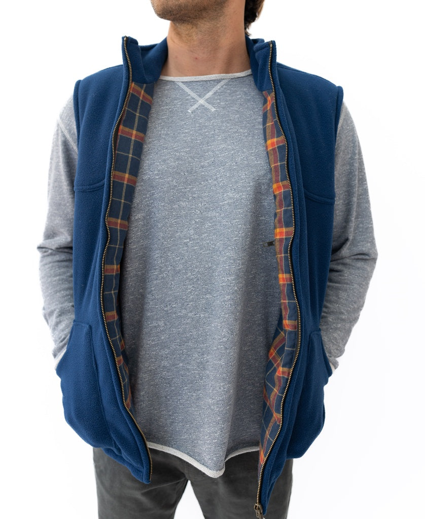 A man wears the Golden Breed Thredbo Vest | Midnight—a blue polyester polar fleece vest with plaid lining—over a gray long-sleeve shirt and dark pants, shown from the shoulders down against a white background.