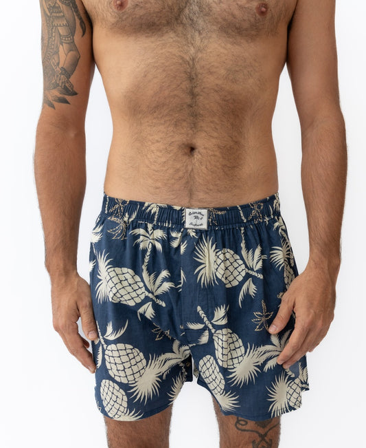 A man wears Golden Breeds Tyson Boxer Short | Vintage Navy, featuring a beige pineapple print, against a white background. Made from 100% cotton, these boxers combine comfort with playful style.