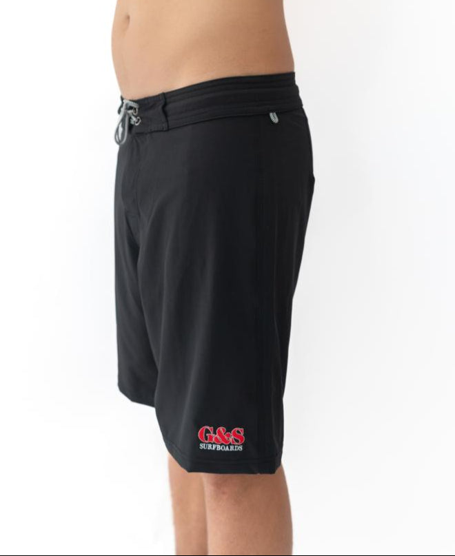 A person wears Gordon & Smith’s G&S Boardie | Black shorts, featuring a drawstring waist and red G&S Surfboards logo on the left leg, made from recycled polyester. They stand against a plain white background.