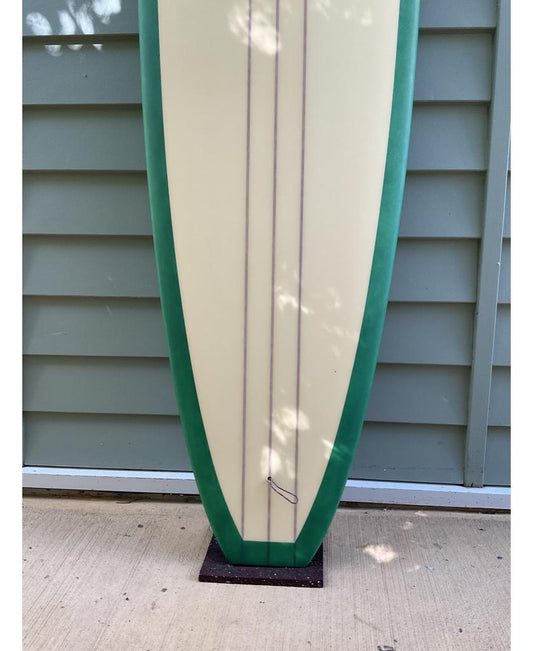 The Gordon & Smith G&S 96 Easy Rider Triple Stringer in Green/Cream stands upright on concrete, showcasing its user-friendly design, triple stringers, and green edges against light green siding.
