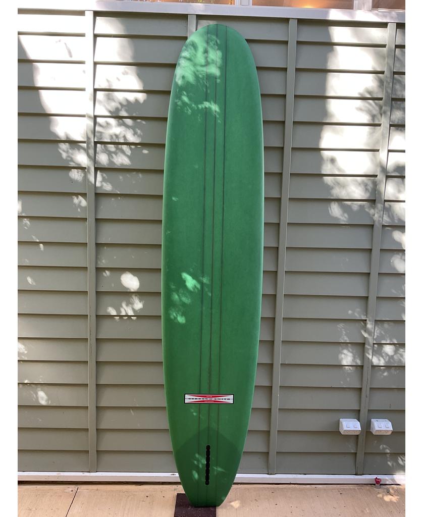 The Gordon & Smith G&S 96 Easy Rider Triple Stringer in Green/Cream stands upright against a light green paneled fence, its user-friendly longboard shape complemented by soft tree shadows playing across the panels.