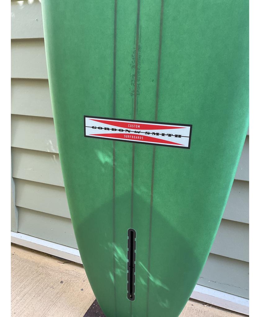 The G&S 96 Easy Rider Triple Stringer by Gordon & Smith, in green/cream with a fin box slot, leans against a light-colored wall.