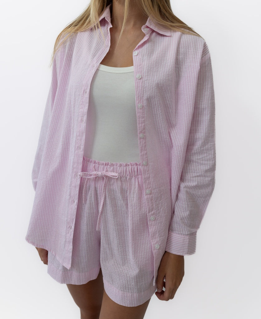 A person wears the Lazy Beach Maeve L/S Shirt in Baby Pink Stripe, styled over a white tank and matching shorts, standing against a plain white background.