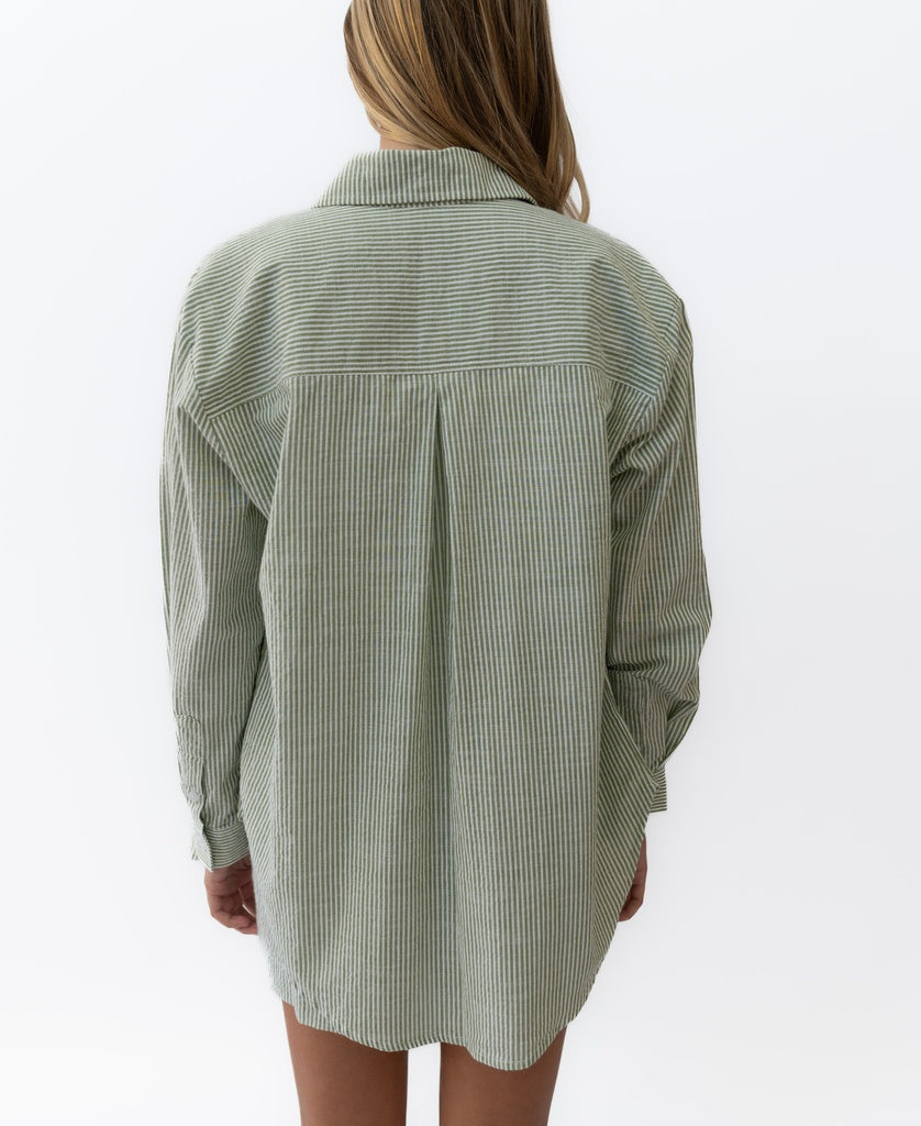 A person with long hair is shown from the back wearing the Lazy Beach Maeve L/S Shirt in Olive Stripe cotton seersucker against a white background.