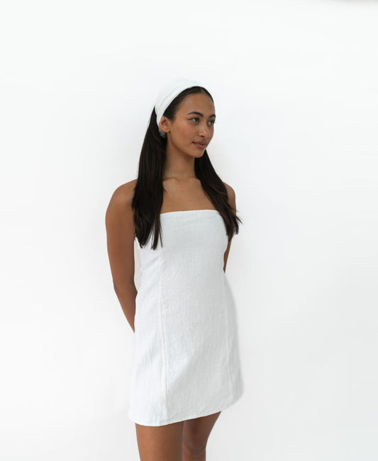 A woman with long dark hair wears the Lazy Beach Head Scarf in White and a strapless white dress, standing against a plain white background with her hands behind her back.