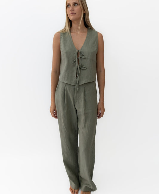 A woman stands barefoot against a white background wearing Lazy Beach’s Janine Pant Linen in dark khaki—an oversized, structured, sleeveless linen two-piece featuring a tie-front top and wide-leg pants.