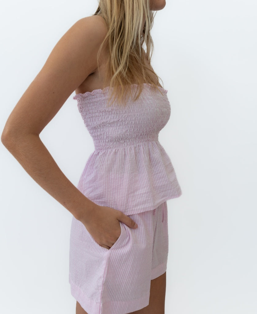 A woman wears the Lazy Beach Judith Short in Baby Pink Stripe with a matching strapless, smocked cotton seersucker top, standing against a plain white background.
