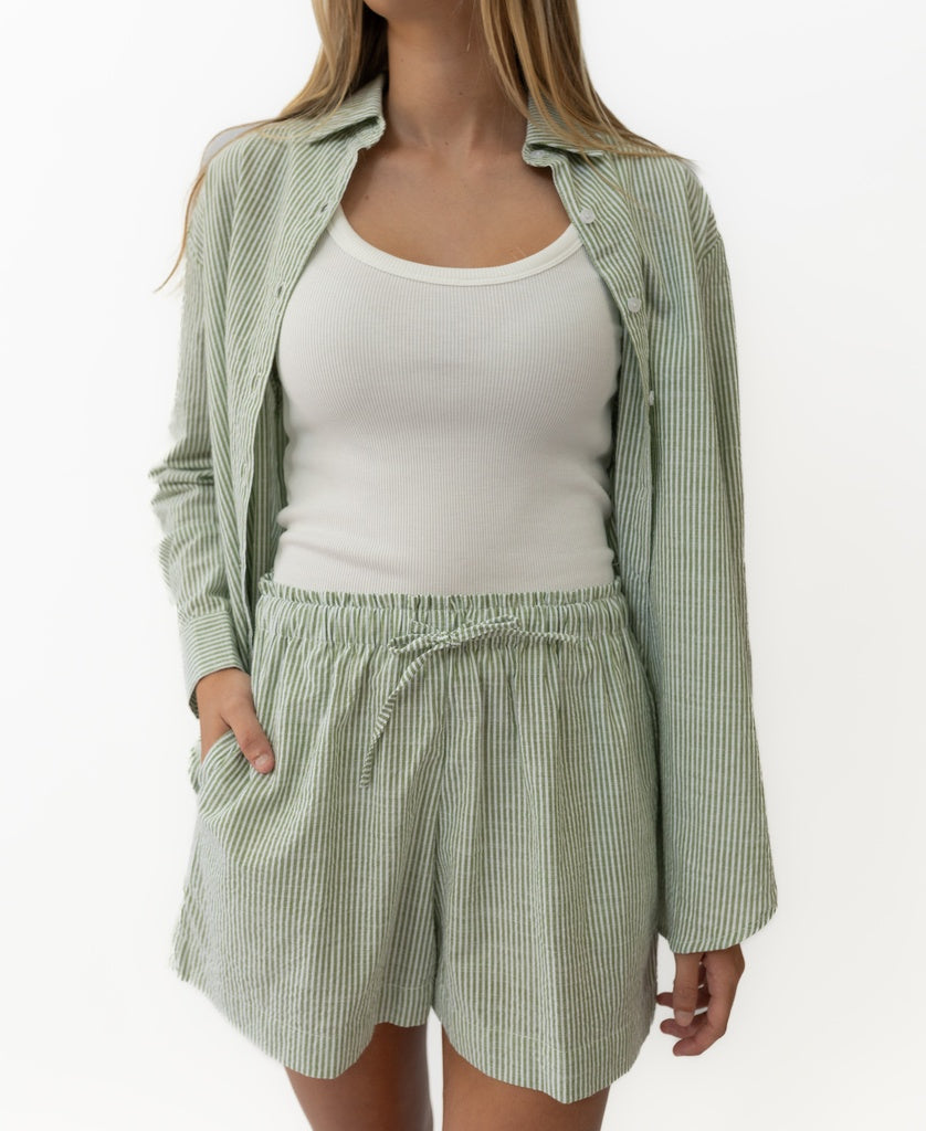 Wearing the Lazy Beach Judith Short in Olive Stripe—handmade in Indonesia with an elastic waist—and a matching cotton button-up shirt, layered over a white ribbed tank top, with one hand in their pocket.