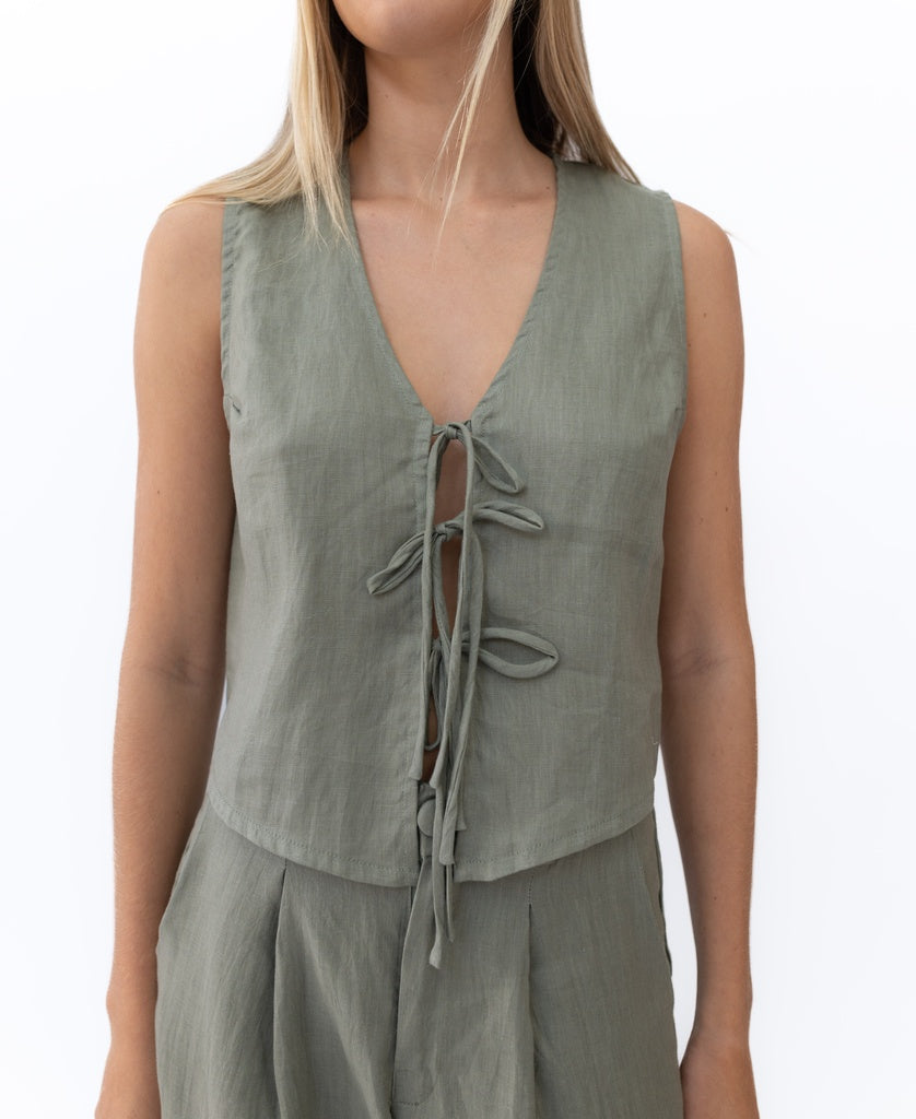 A person wears the Lazy Beach Rennie Tie Vest Linen in Dark Khaki, a sleeveless vest with a deep V-neck and three front ties, paired with matching high-waisted pants, all handmade in Bali.