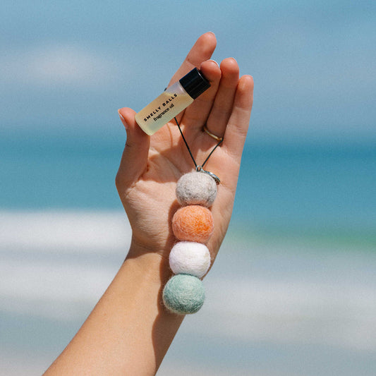 A hand holds a vial labeled Smelly Balls Seapink Set Sweetpea and a string of four colorful, ethically sourced wool felt balls against a blurred beach background.
