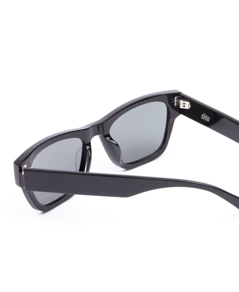 The Sito Boston | Black / Iron Grey Polar sunglasses feature a stylish black rectangular plant-based acetate frame with dark lenses for high UV protection. Shown angled on a white background, they blend sustainability and modern design.