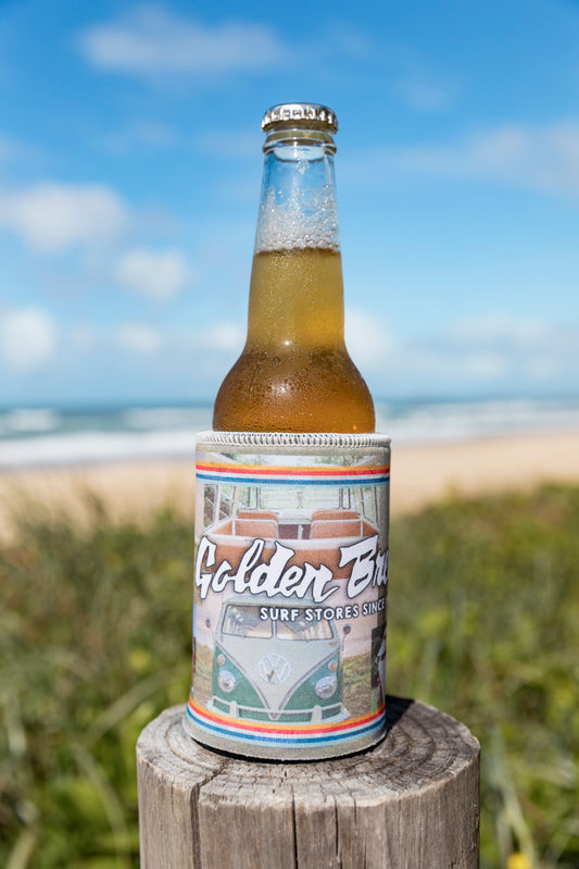 A cold beer in the limited edition Golden Breed GB Kombi Collage Stubby sits on a wooden post, overlooking the beach and ocean in the background.