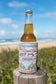 A cold beer in the limited edition Golden Breed GB Kombi Collage Stubby sits on a wooden post, overlooking the beach and ocean in the background.