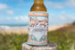 A GB Kombi Collage Stubby by Golden Breed, featuring a colorful Kombi design, holds a beer bottle on a wooden post by the beach.