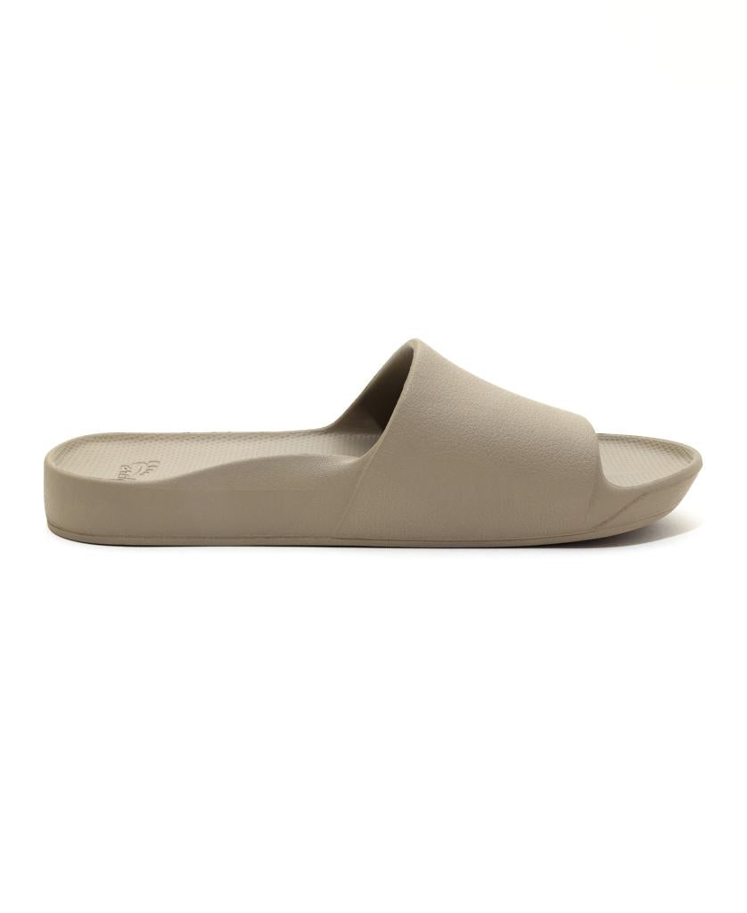 The Archies Archie Slide in Taupe is a beige slip-on sandal with a thick sole and wide upper strap, made from closed cell foam that molds to your foot, shown side-on against a plain white background.