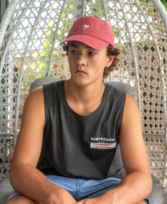 A young person in a sleeveless black shirt, blue shorts, and a Golden Breed Palm Cap in Wash Red sits on a wicker chair indoors, looking to the side.