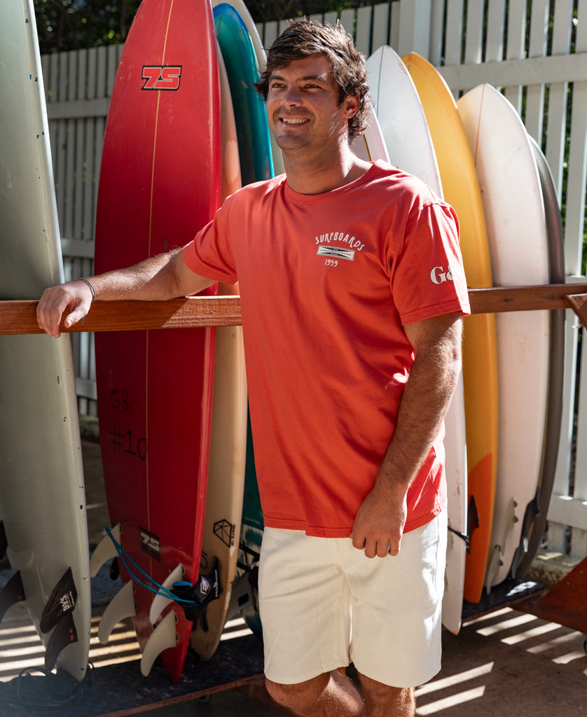 A man wearing the Gordon & Smith G&S Surfboards Tee in Wash Red and white shorts stands by a rack of surfboards, smiling as sunlight casts shadows on the scene.